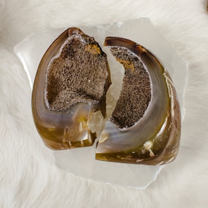 May include: A brown and white agate geode with a natural crack, revealing the inner crystal formations. The geode is displayed on a white selenite slab.