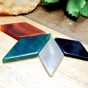 May include: Four diamond-shaped agate stones in different colors: red, green, white, and black. The stones are arranged in a star shape on a wooden surface.