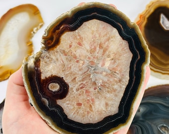 Pode incluir: Uma fatia de ágata polida com um centro castanho claro, rodeada por uma borda castanha escura e preta. A pedra natural tem uma forma circular e um padrão único. Outras fatias de ágata são visíveis no fundo.