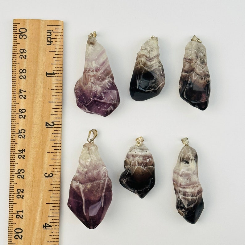 May include: Six amethyst crystal pendants with gold-tone bail settings. The crystals are various shades of purple and white, with some having a black or dark purple hue.