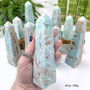 May include: A collection of blue and white banded aragonite crystal points, also known as "Angel Aura" aragonite. The points are arranged in a row, with one point being held in a hand. The points are approximately 401 to 500 grams in weight.