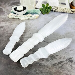May include: Three white selenite wands of varying sizes, with a pointed tip and a textured, twisted handle. The wands are arranged on a gray surface.