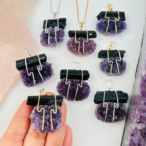May include: Six black tourmaline and amethyst crystal pendants with gold and silver wire wrapping. The pendants are displayed on a white surface.