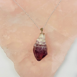 May include: A silver chain necklace with a purple and white crystal pendant. The pendant is a natural, rough-cut crystal with a unique shape.