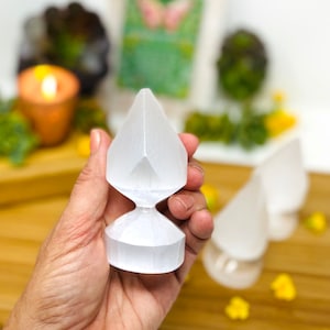May include: A white selenite crystal wand with a faceted, teardrop-shaped top and a cylindrical base. The wand is held in a hand, with two similar wands in the background. The background includes a lit candle and greenery.