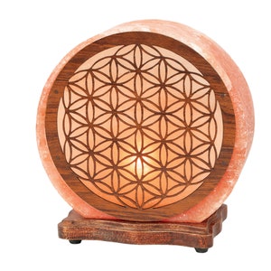 K&ouml;nnte beinhalten: Eine runde Himalaya-Salzlampe mit einem Holzsockel und einem geschnitzten Lebensblume-Design. Die Lampe ist beleuchtet und leuchtet in einem warmen Orangeton.