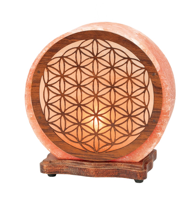 K&ouml;nnte beinhalten: Eine runde Himalaya-Salzlampe mit einem Holzsockel und einem geschnitzten Lebensblume-Design. Die Lampe ist beleuchtet und leuchtet in einem warmen Orangeton.