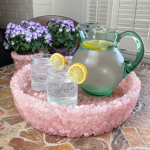 May include: A pink rose quartz tray with a green glass pitcher of lemonade and two glasses of lemonade with lemon slices. The tray is filled with small rose quartz stones.