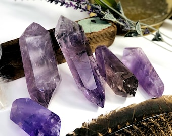 May include: Six amethyst crystal points in various sizes and shades of purple, arranged on a white surface with a feather and a wooden board.