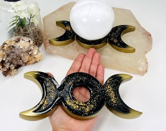 May include: Black and gold triple moon altar stand with a glitter finish. The stand is made of resin and is perfect for holding crystals, candles, or other altar items.