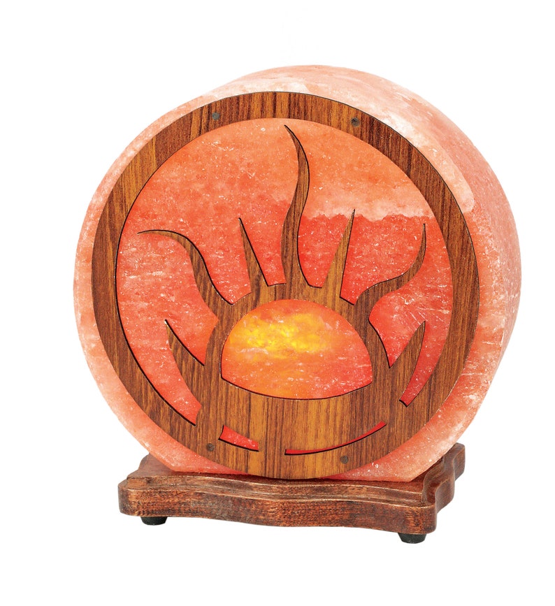 K&ouml;nnte beinhalten: Eine runde Himalaya-Salzlampe mit einem Holzrahmen, der ein geschnitztes Sonnenmotiv zeigt. Die Lampe ist eingeschaltet und leuchtet orange.
