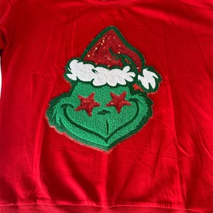 May include: Red Christmas sweater with a green, sequined Grinch wearing a Santa hat with red and white trim. The Grinch's eyes are red stars.