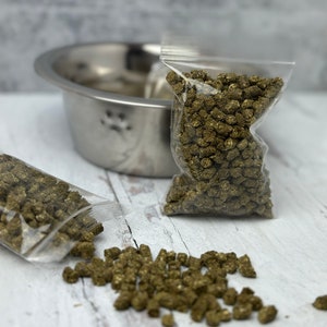 May include: A stainless steel pet bowl with paw print detail, next to two clear plastic bags filled with green pet food pellets. Some pellets are scattered on a white wooden surface. The food is for small animals.