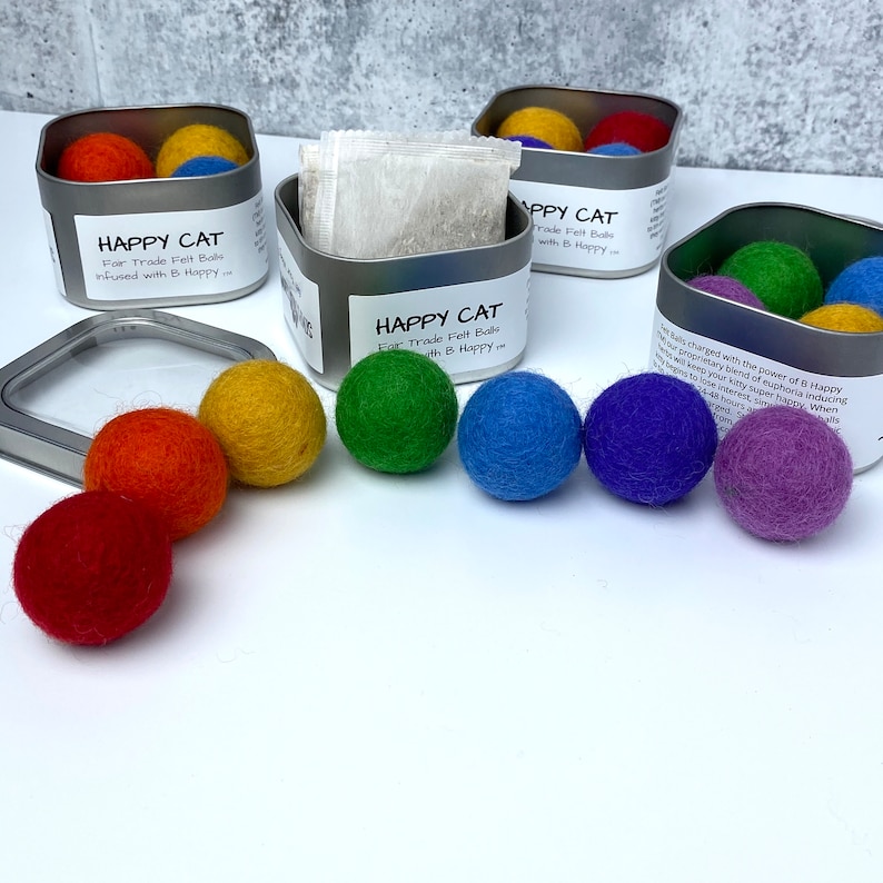 Pu&ograve; includere: Un set di palline di feltro colorate in un motivo arcobaleno, confezionate in una scatola di latta argentata con il testo "HAPPY CAT" e "For Trade Felt Balls infused with B Happy".