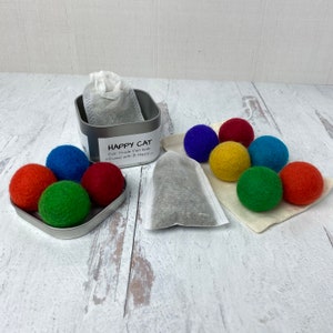 Pu&ograve; includere: Un contenitore di latta pieno di palline di feltro colorate per gatti. La latta ha un'etichetta che recita "Happy Cat Fair Trade Felt Balls infused with B Happy". Le palline di feltro sono anche in un piccolo sacchetto bianco con una bustina di t&egrave; grigia.