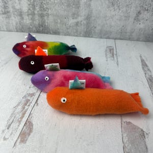May include: Four plush fish toys arranged in a row. The top fish is multi-colored with a tie-dye pattern, the second is dark maroon, the third is pink and purple, and the bottom fish is solid orange. Each fish features a small white eye and a fin.