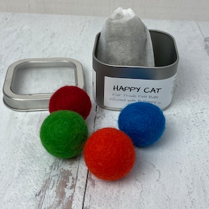 Pu&ograve; includere: Quattro palline di feltro colorate in rosso, verde, arancione e blu, accanto a una scatola di latta argentata con un'etichetta bianca che recita "HAPPY CAT Fair Trade Felt Balls Infused with B. Happy TM".