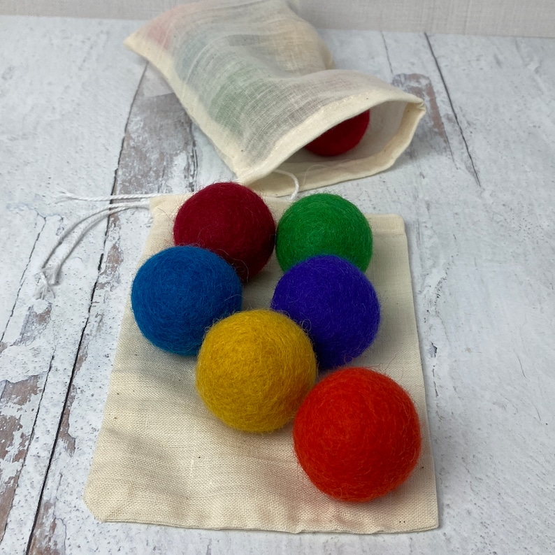 Pu&ograve; includere: Un set di sei palline di feltro colorate in una borsa di cotone bianco con coulisse. Le palline sono rosse, arancioni, gialle, verdi, blu e viola.