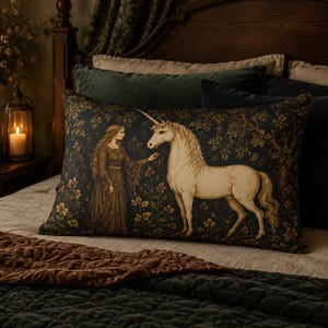 May include: A rectangular throw pillow featuring a dark-toned tapestry-style print of a person in a long gown standing before a white unicorn. The scene is set against a backdrop of dense foliage and flowers, rendered in shades of green and gold.