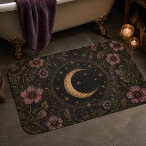 May include: A rectangular bath mat featuring a dark background with a central golden crescent moon surrounded by small stars. The design includes botanical illustrations of pink flowers and green foliage arranged in a circular pattern.