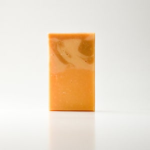 Peut inclure: Un pain de savon rectangulaire aux teintes orange et beige. Le savon a une surface lisse avec un motif marbré sur le dessus. Le savon est posé verticalement sur un fond blanc.