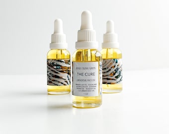 May include: Three glass bottles of "THE CURE" universal face oil with white droppers. The bottles have a yellow liquid inside and are labeled with ingredients and a 1 oz size. The labels have a beige, dusk, and grey pattern.