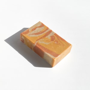 May include: A rectangular bar of handmade soap with a swirled design. The soap features shades of yellow, orange, and tan, creating a marbled effect. The bar is set against a white background, with a shadow cast to the left.