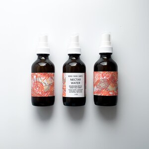 May include: Three brown glass spray bottles with white spray nozzles. The bottles have labels with text and a floral design in shades of red, beige, and gray. The center bottle's label reads "Nectar Water" and "Nourishing Rose & Honey Face Toner."