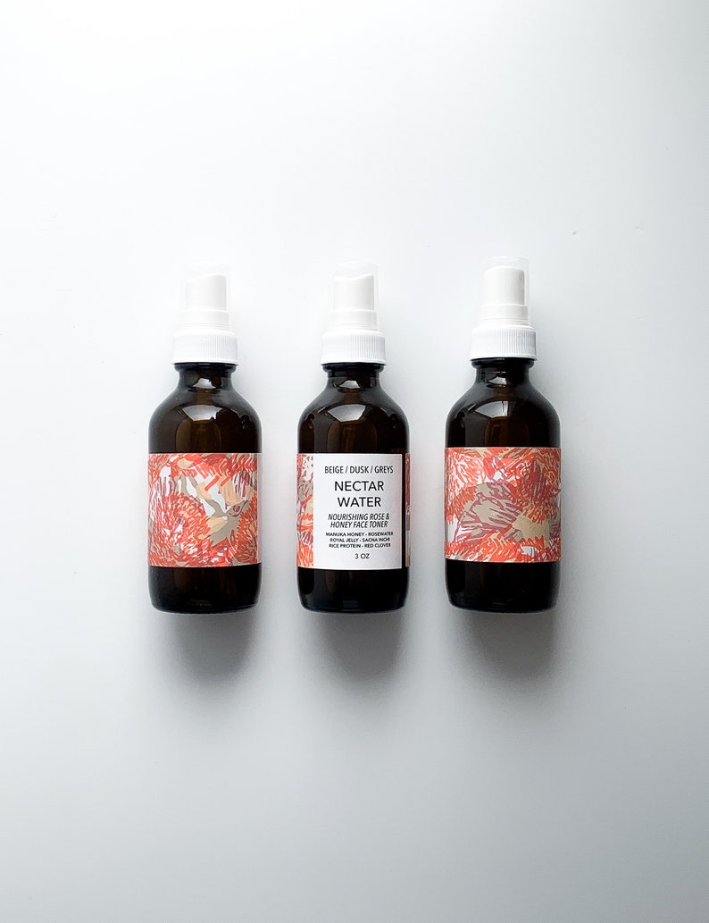 May include: Three brown glass spray bottles with white spray nozzles. The bottles have labels with text and a floral design in shades of red, beige, and gray. The center bottle's label reads "Nectar Water" and "Nourishing Rose & Honey Face Toner."