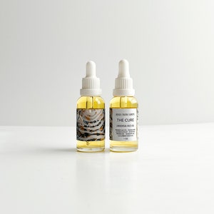 May include: Two glass bottles of face oil with white droppers. The bottles are filled with a yellow liquid and have labels with text that reads "THE CURE UNIVERSAL FACE OIL". The labels also list ingredients and the size, 1 oz.