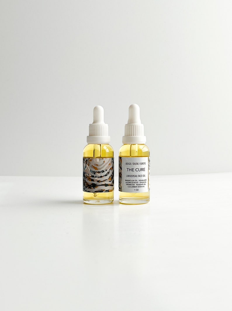 May include: Two glass bottles of face oil with white droppers. The bottles are filled with a yellow liquid and have labels with text that reads "THE CURE UNIVERSAL FACE OIL". The labels also list ingredients and the size, 1 oz.