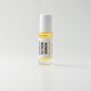 Puede incluir: Una botella de vidrio transparente de Nomad Mani Oil con tapa blanca. La botella contiene un l&iacute;quido amarillo y tiene una etiqueta con el texto "Nomad Mani Oil" y otro texto. Es un tratamiento para cut&iacute;culas con manteca de mango y aceite de jojoba.