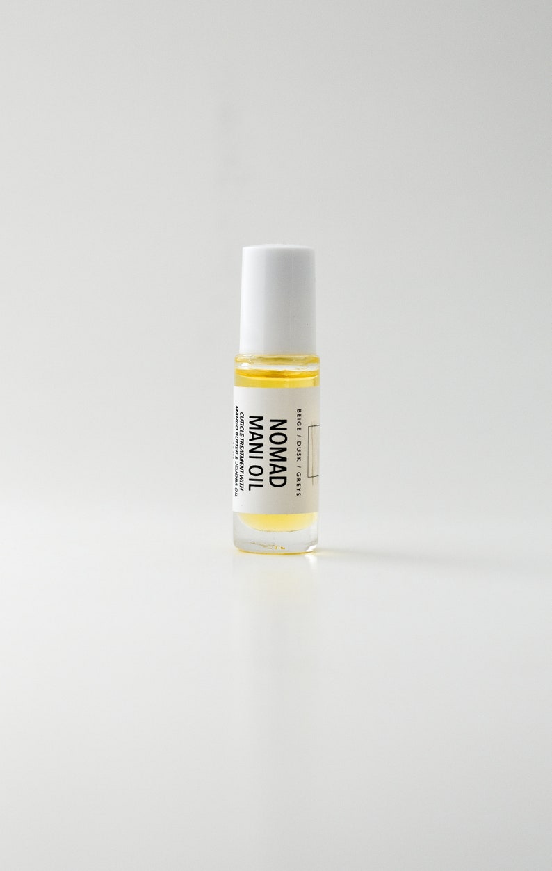 Puede incluir: Una botella de vidrio transparente de Nomad Mani Oil con tapa blanca. La botella contiene un l&iacute;quido amarillo y tiene una etiqueta con el texto "Nomad Mani Oil" y otro texto. Es un tratamiento para cut&iacute;culas con manteca de mango y aceite de jojoba.