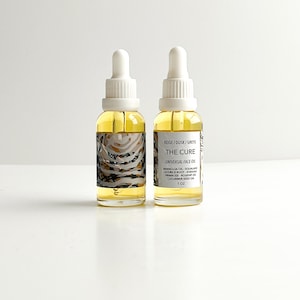 May include: Two glass bottles of "THE CURE" universal face oil with white droppers. The labels feature text and a beige, gray, and black pattern. The bottles contain a yellow liquid and are labeled "1 oz".