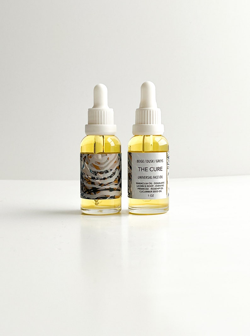 May include: Two glass bottles of "THE CURE" universal face oil with white droppers. The labels feature text and a beige, gray, and black pattern. The bottles contain a yellow liquid and are labeled "1 oz".