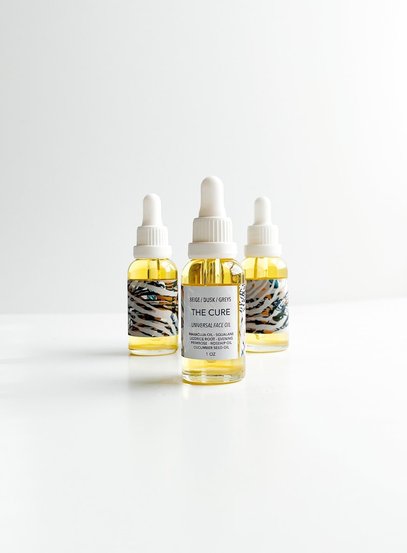 May include: Three glass bottles of "THE CURE" universal face oil with white droppers. The bottles have a yellow liquid inside and are labeled with ingredients and a 1 oz size. The labels have a beige, dusk, and grey pattern.