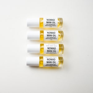 Puede incluir: Cuatro botellas de vidrio transparente de Nomad Mani Oil, cada una con una tapa blanca y una etiqueta. Las etiquetas dicen "Nomad Mani Oil" y enumeran los ingredientes. Las botellas est&aacute;n llenas de un l&iacute;quido amarillo.