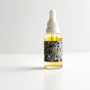 May include: A clear glass bottle with a white dropper top, filled with a yellow liquid. The bottle has a decorative label with a gray background and colorful abstract shapes. The bottle is likely a skincare product.