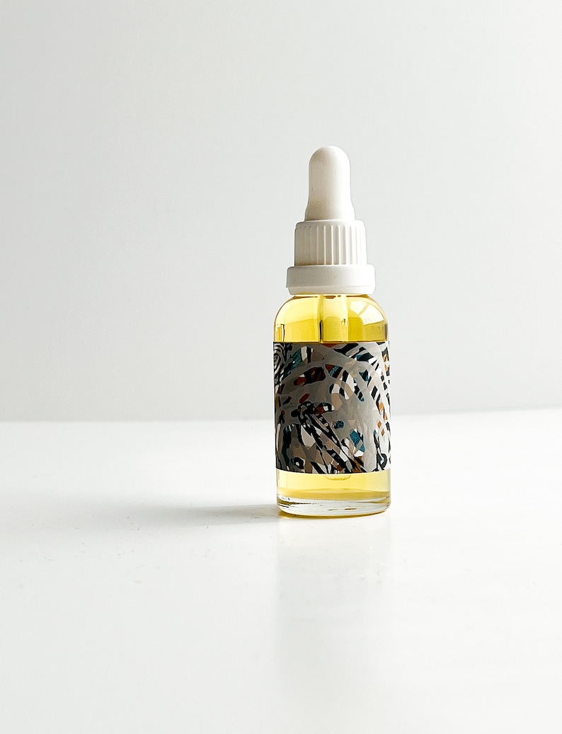 May include: A clear glass bottle with a white dropper top, filled with a yellow liquid. The bottle has a decorative label with a gray background and colorful abstract shapes. The bottle is likely a skincare product.