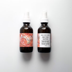 May include: Two brown glass spray bottles with white spray nozzles. The bottles have labels with text and a floral design in red, beige, and white. The text on the label reads "NECTAR WATER" and "NOURISHING ROSE & HONEY FACE TONER". Each bottle contains 3 oz.