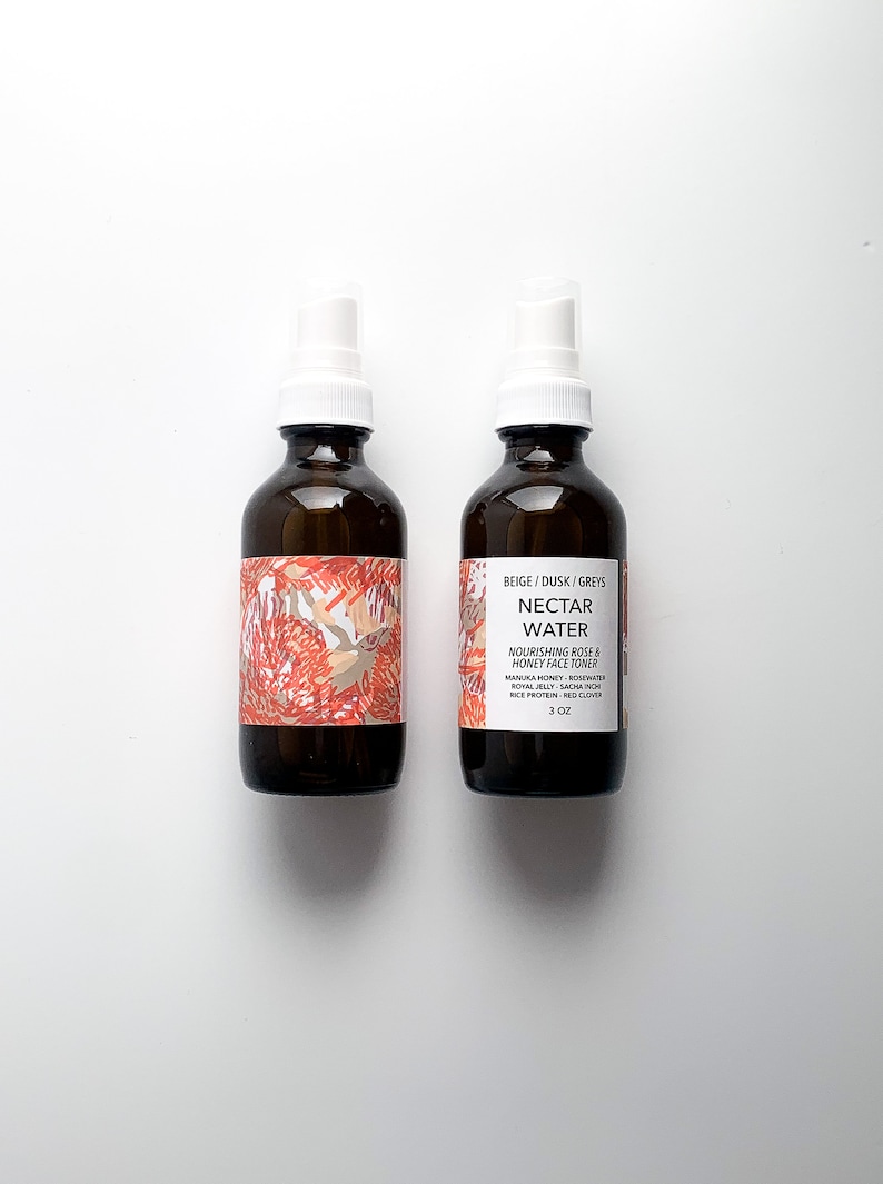 May include: Two brown glass spray bottles with white spray nozzles. The bottles have labels with text and a floral design in red, beige, and white. The text on the label reads "NECTAR WATER" and "NOURISHING ROSE & HONEY FACE TONER". Each bottle contains 3 oz.