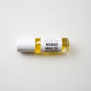 Puede incluir: Un peque&ntilde;o frasco de vidrio transparente con un l&iacute;quido amarillo, etiquetado como "NOMAD MANI OIL". El frasco tiene una tapa blanca y una etiqueta con texto negro. Es un tratamiento para cut&iacute;culas con manteca de mango y aceite de jojoba.