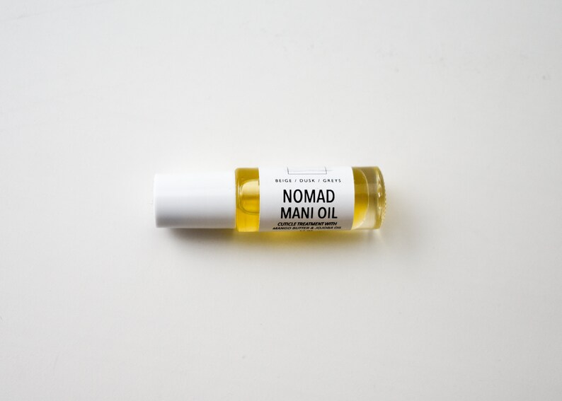 Puede incluir: Un peque&ntilde;o frasco de vidrio transparente con un l&iacute;quido amarillo, etiquetado como "NOMAD MANI OIL". El frasco tiene una tapa blanca y una etiqueta con texto negro. Es un tratamiento para cut&iacute;culas con manteca de mango y aceite de jojoba.