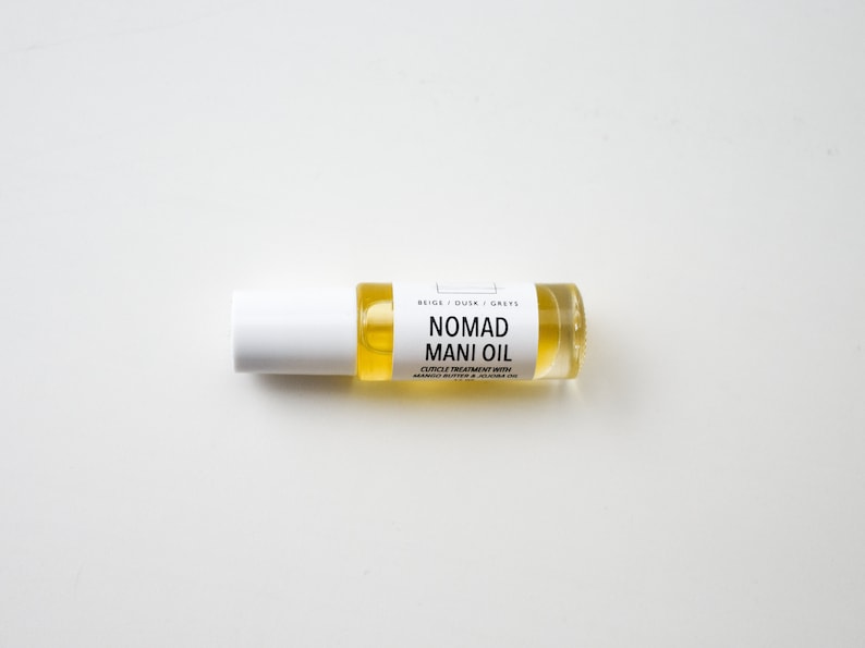 Puede incluir: Una peque&ntilde;a botella de vidrio transparente de Nomad Mani Oil con una tapa blanca. La botella contiene un l&iacute;quido amarillo y tiene una etiqueta blanca con texto negro que dice "NOMAD MANI OIL". La etiqueta tambi&eacute;n incluye el texto "CUTICLE TREATMENT WITH MANGO BUTTER & JOJOBA OIL".
