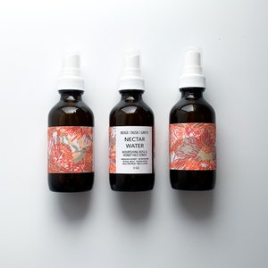 May include: Three brown glass bottles with white spray tops. Each bottle has a label with a floral design in shades of orange, beige, and gray. The center bottle's label reads "Nectar Water" and lists ingredients.