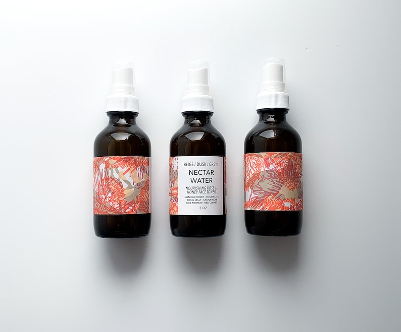 May include: Three brown glass bottles with white spray tops. Each bottle has a label with a floral design in shades of orange, beige, and gray. The center bottle's label reads "Nectar Water" and lists ingredients.