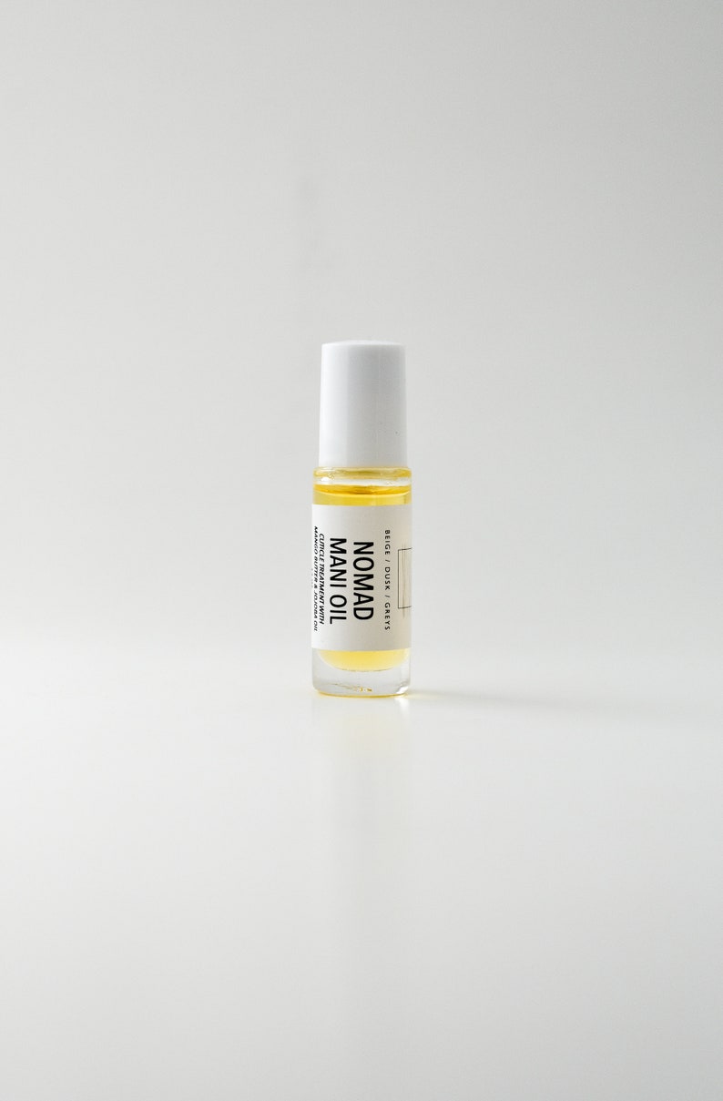 Puede incluir: Una botella de vidrio transparente de Nomad Mani Oil con tapa blanca. La botella contiene un l&iacute;quido amarillo y tiene una etiqueta con texto negro. El producto est&aacute; dise&ntilde;ado para el tratamiento de cut&iacute;culas con manteca de marula y aceite de jojoba.
