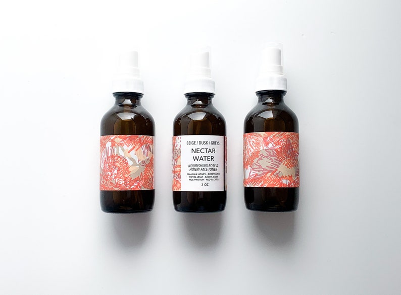 May include: Three brown glass bottles with white spray tops. Each bottle has a label with a floral design in shades of red, beige, and gray. The center bottle's label reads "Nectar Water" and "Nourishing Rose & Honey Face Toner", with a 3 oz size.