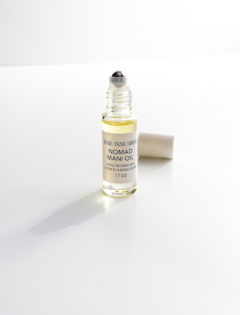 Puede incluir: Un frasco roll-on de vidrio transparente lleno de l&iacute;quido amarillo, etiquetado "BEIGE / DUSK / GREYS NOMAD MANI OIL". El frasco tiene una bola rodante plateada y una tapa blanca. La etiqueta tambi&eacute;n menciona tratamiento de cut&iacute;culas. Contiene 5 ml.