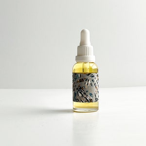 May include: A clear glass bottle with a white dropper top, filled with a yellow liquid. The bottle has a decorative label with a pattern of leaves in shades of gray, blue, and brown. The bottle is sitting on a white surface.